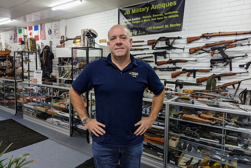 A middle-aged man stands with his hands on his hips in front of a wall of antique rifles.