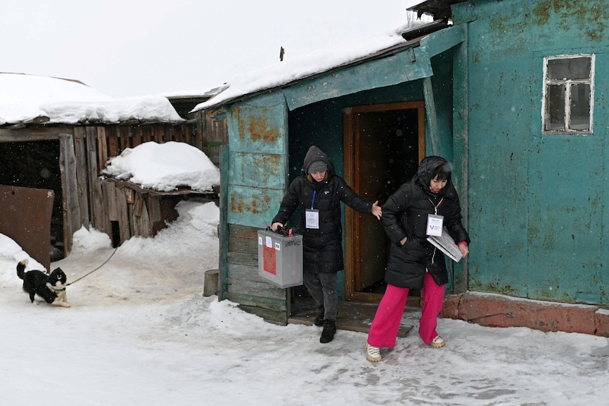 Two women, one carrying a ballot box and the other holding a clipboard, walk outdoors in the snow near houses