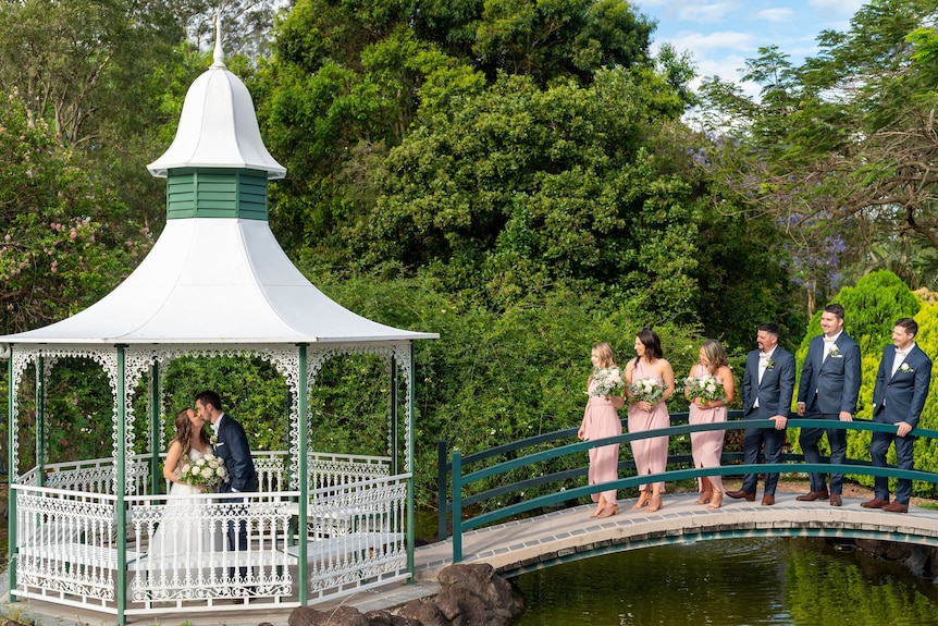 an image of a bride and groom kissing in a rotunda with the bridal party standing on a bridge leading away from rotunda,  