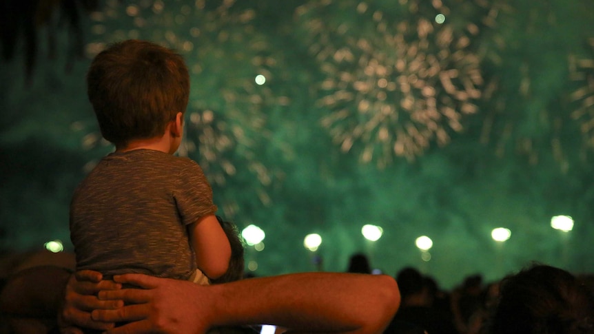 A boy sits on a man's shoulders watching a fireworks display.