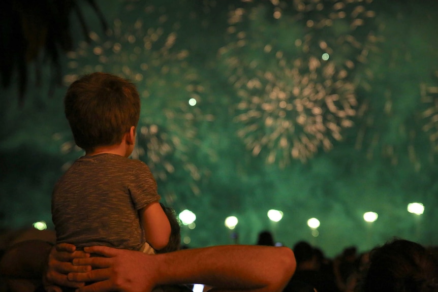 A boy sits on a man's shoulders watching a fireworks display.