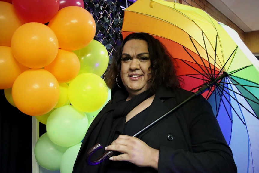 A man stands holding a rainbow umbrella in front of rainbow coloured balloons. 