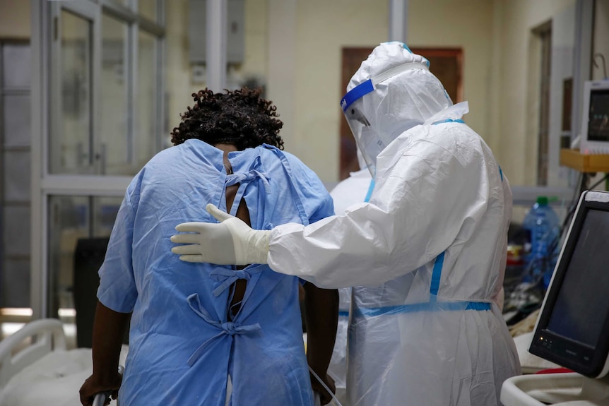 A doctor wearing a white PPE suit helps a patient with a metal walker wearing a blue hospital gown.