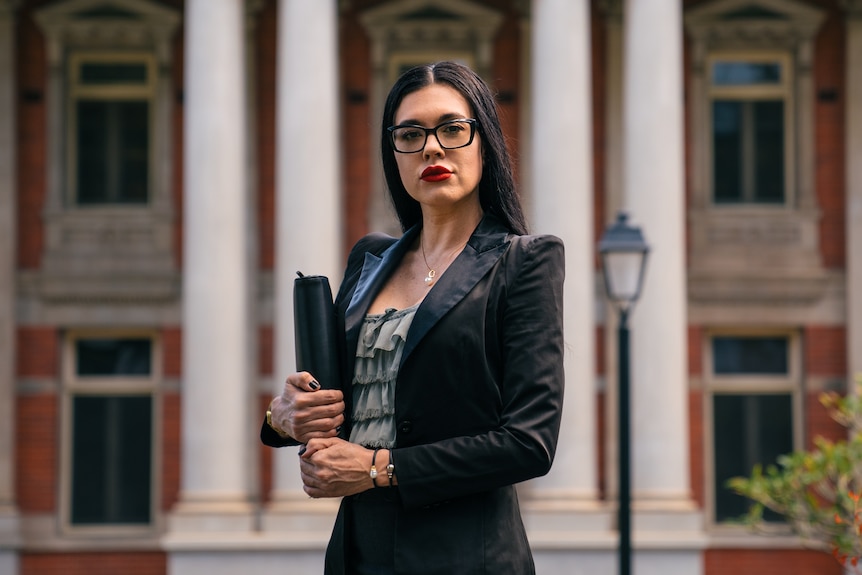 A woman with long black hair stands in front of a building holding a compendium. 