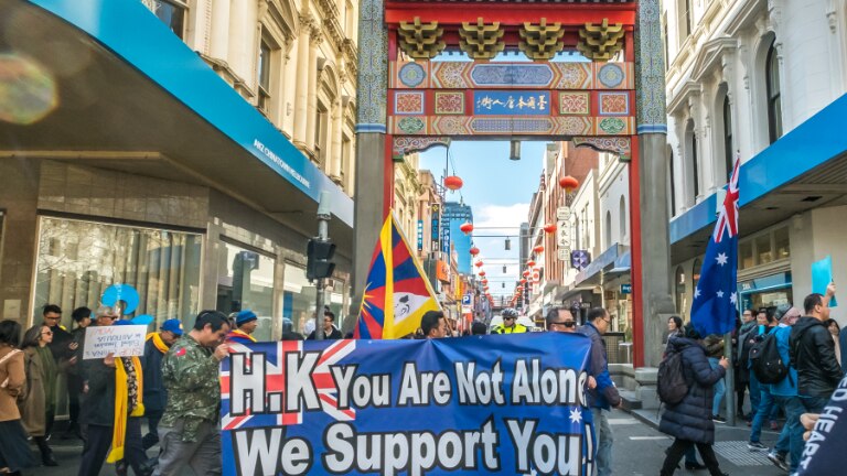Looking down a narrow Melbourne street, you view a crowd of people holding anti-China banners and Australian flags.