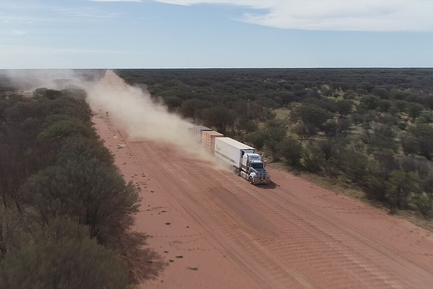 A road train travels along a dirt highway, with dust in its wake.