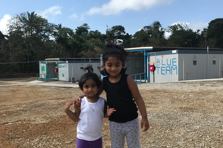 Two young Tamil girls stand in a bare gravel field, two buildings in the background