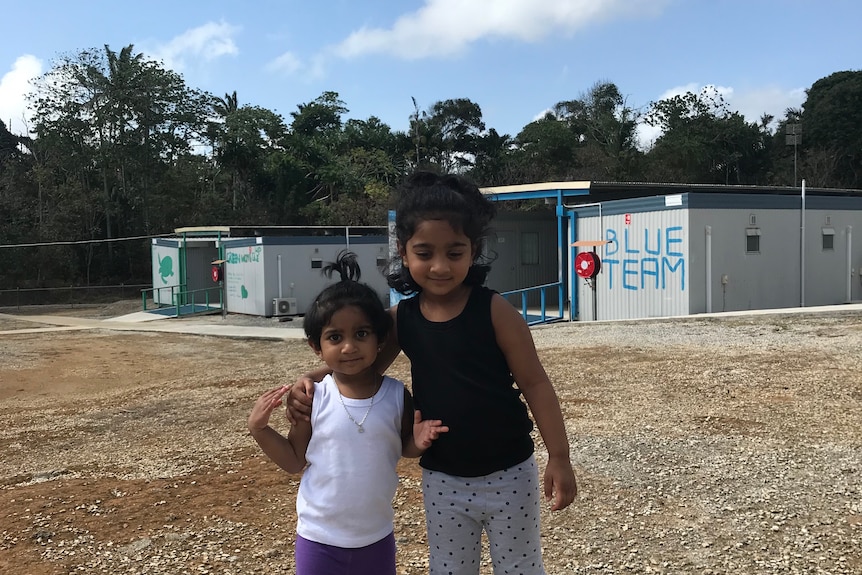 Two young Tamil girls stand in a bare gravel field, two buildings in the background