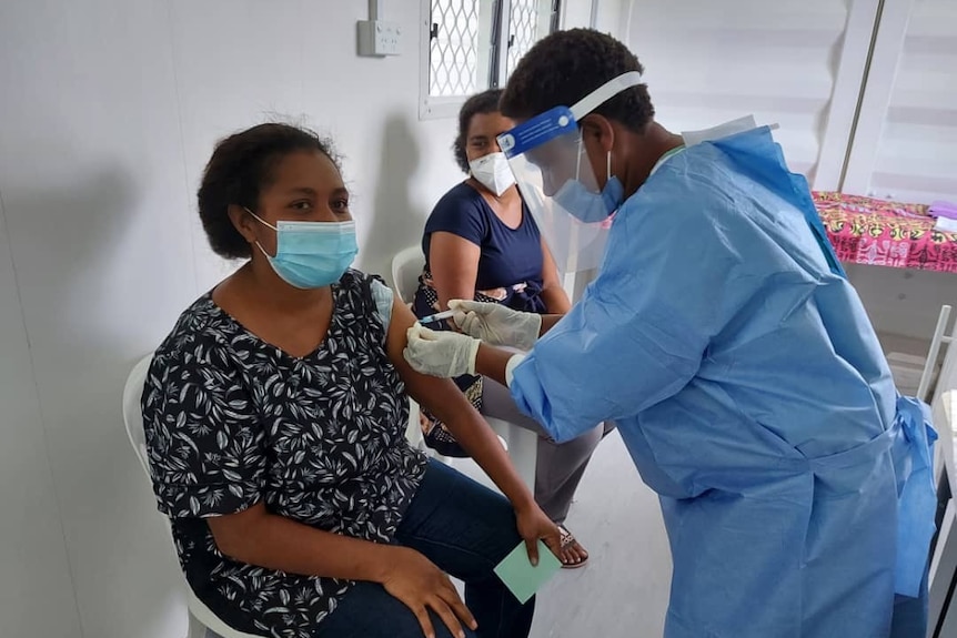 A nurse in full protective gear uses a syringe to innoculate a woman who is sitting in a chair wearing a face mask.