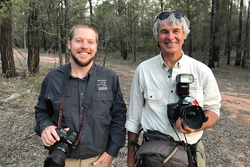 Two men with DSLR cameras around their necks stand in a dry forest.