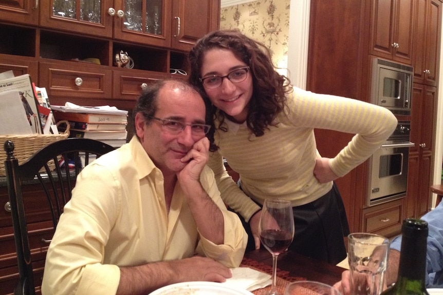 A young woman in glasses smiles stands next to her father in the kitchen