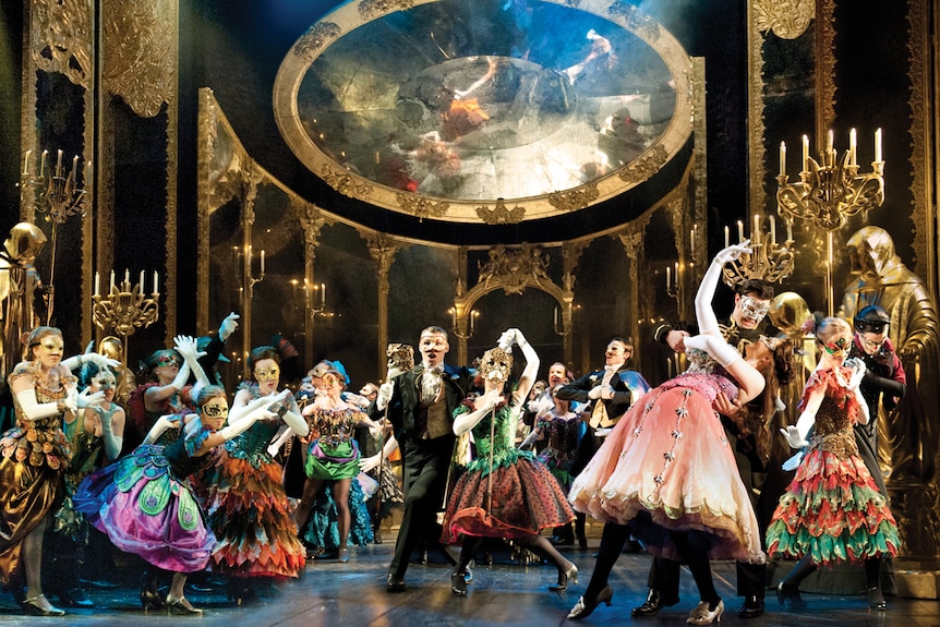 A large cast of opera singers dressed in elaborate costumes perform a masquerade scene from Phantom of the Opera.