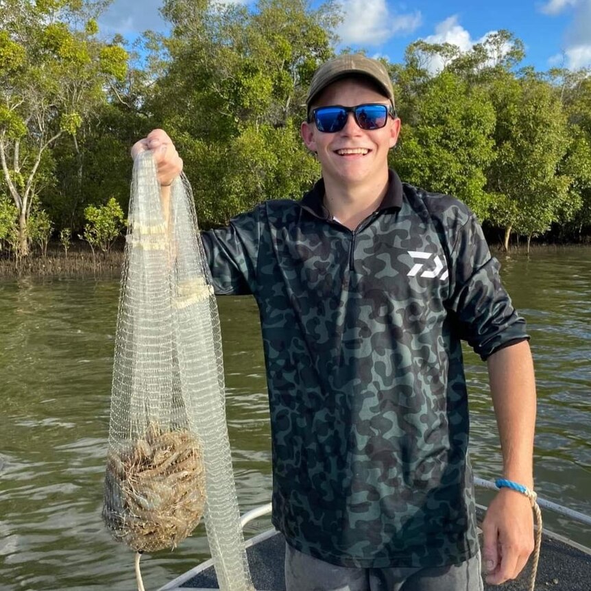 A young man stands in a grey camouflage shirt, green hat and sunglasses holding a cast net full of prawns  in his right and