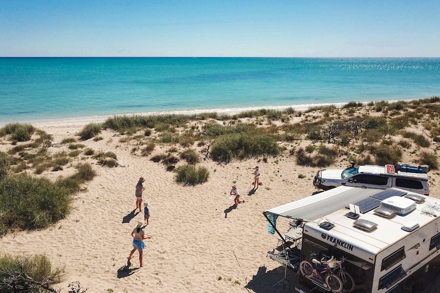 Aerial view of kids running on the sand near a caravan, a few metres from the clear, blue ocean.