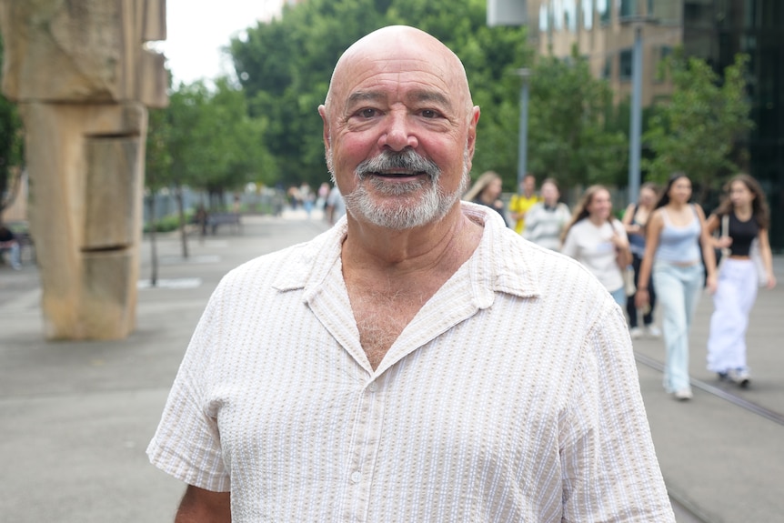 A bald man wearing an open necked shirt smiles while standing on a city street