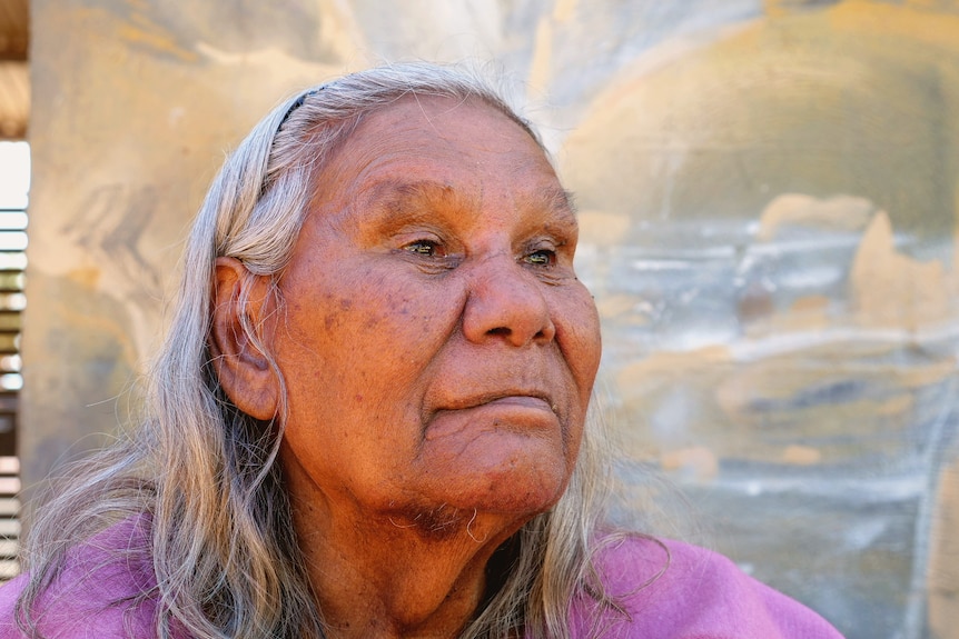 A close -up of a serious grey-haired Indigenous elderly woman, pink top, sits in front of a pale mural.