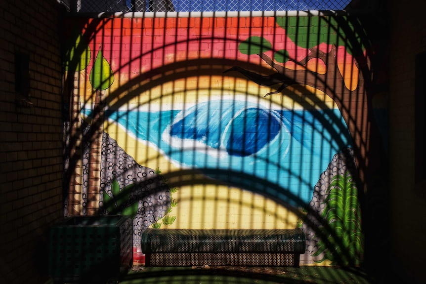 A mural on a brick wall in an outdoor area, the shadows of bars above can be seen on the wall.