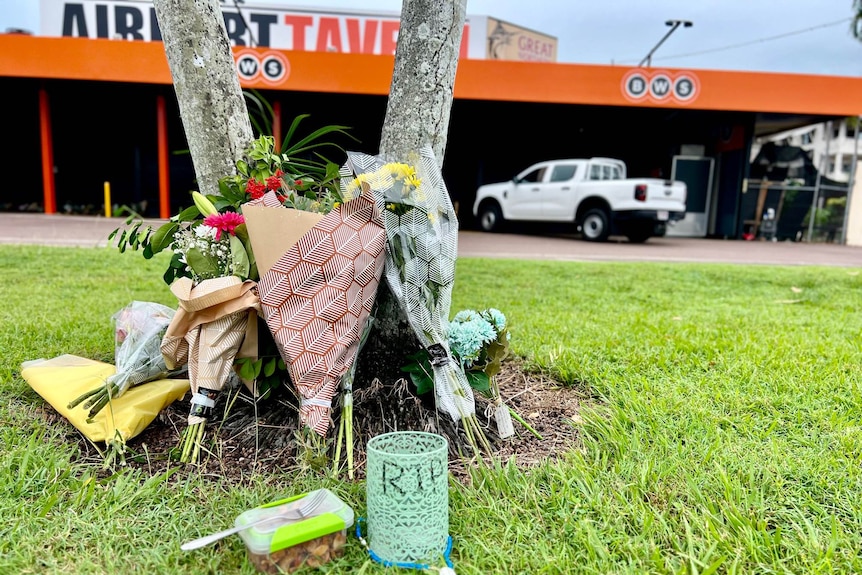 Several bunches of flowers lying on a patch of a grass in front of a bottle shop and car park.