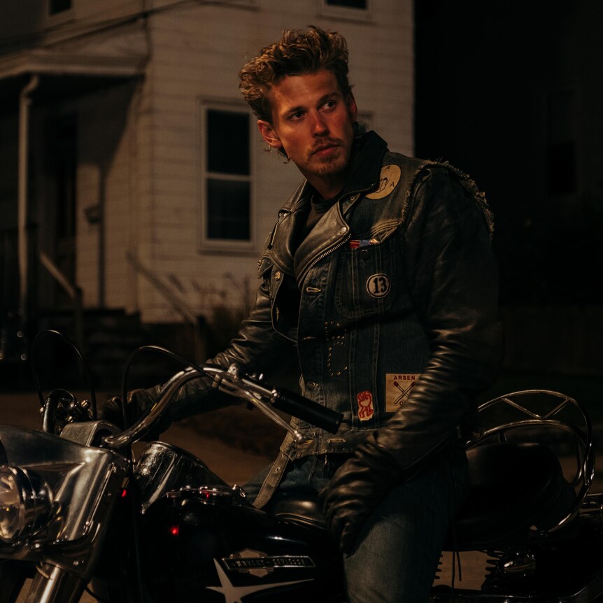 A man in a leather jacket sits on a motorcycle at night staring off camera