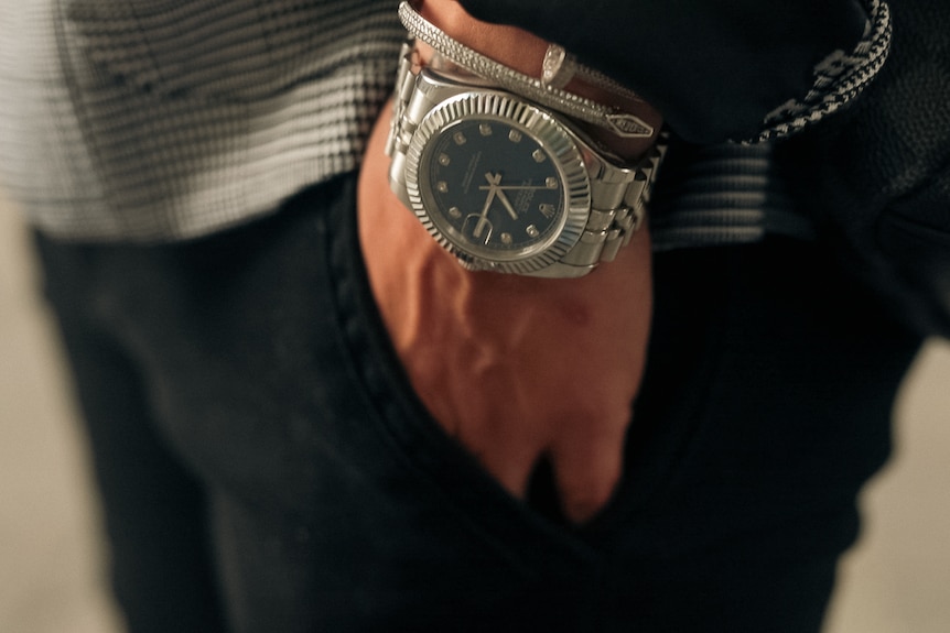 A silver rolex watch on the wrist of a person with their hand in the pocket of black pants.