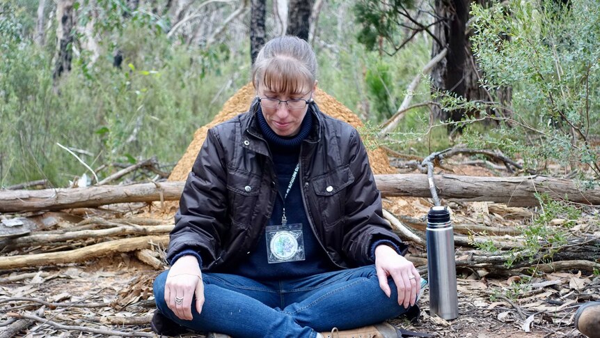 A woman sits cross-logged in a green forest with a thermos of tea by her side