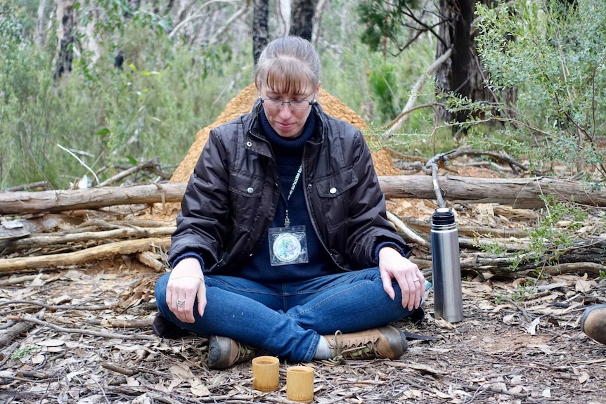 A woman sits cross-logged in a green forest with a thermos of tea by her side