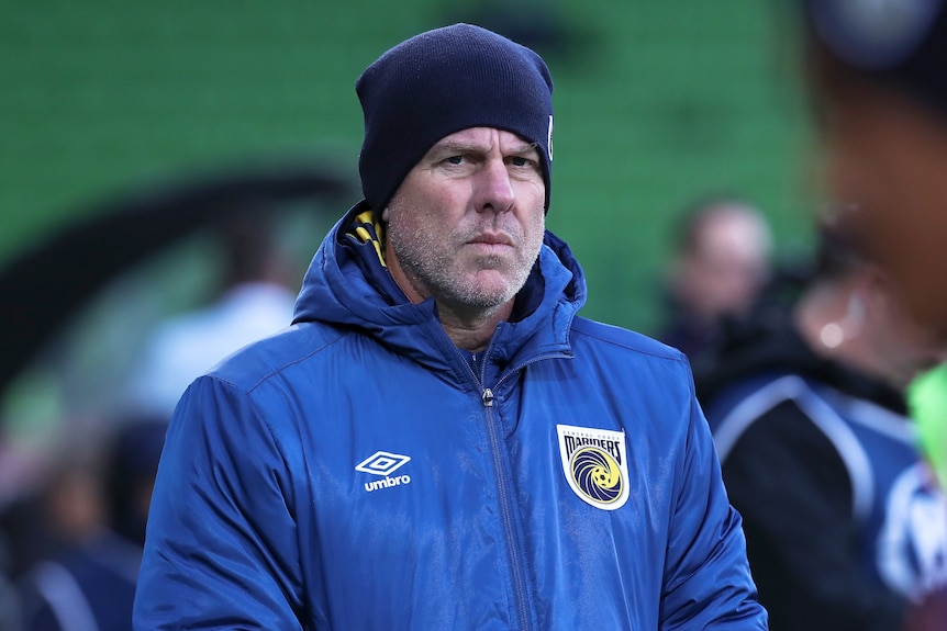 A man wearing a football club jacket and beanie stands looking serious. 