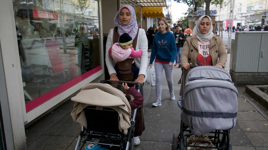 Two women in headscarves push prams down a shopping strip