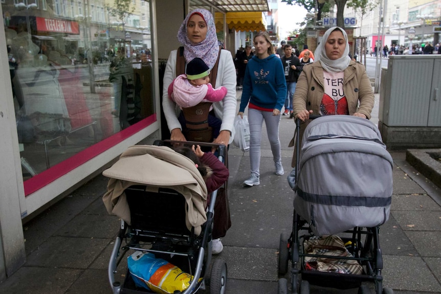 Two women in headscarves push prams down a shopping strip