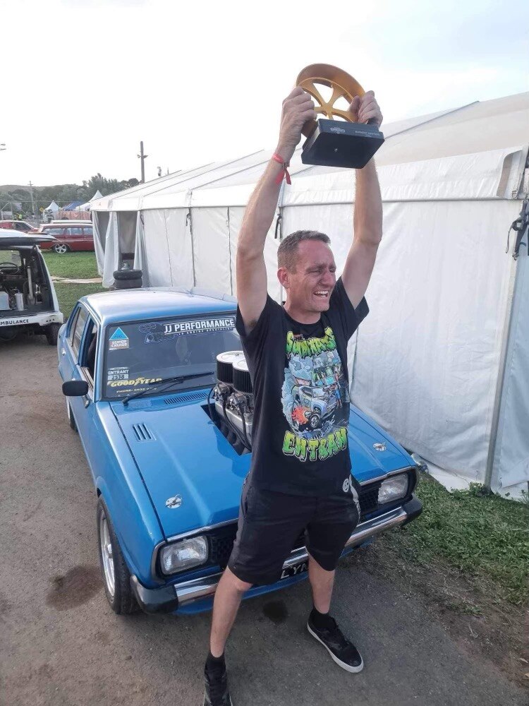 A man stands in front of a blue car with arms outstretched holding a trophy 