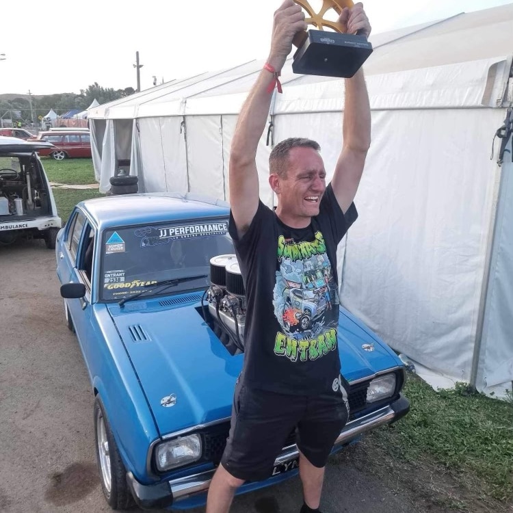 A man stands in front of a blue car with arms outstretched holding a trophy 