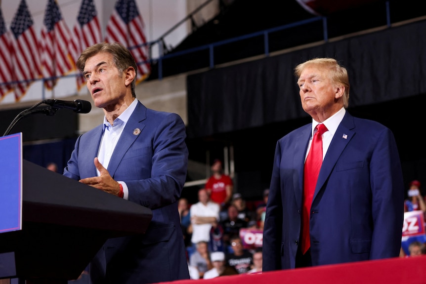 Dr Oz stand behind a podium in suit with Trump looking stern behind him