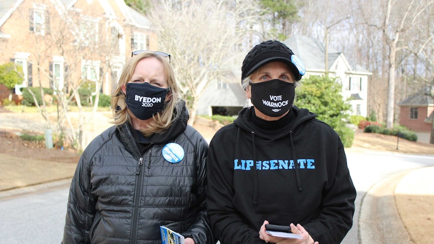 A woman with blonde hair and wearing a Byedon 2020 mask stands next to a woman wearing a black cap and wearing the same mask