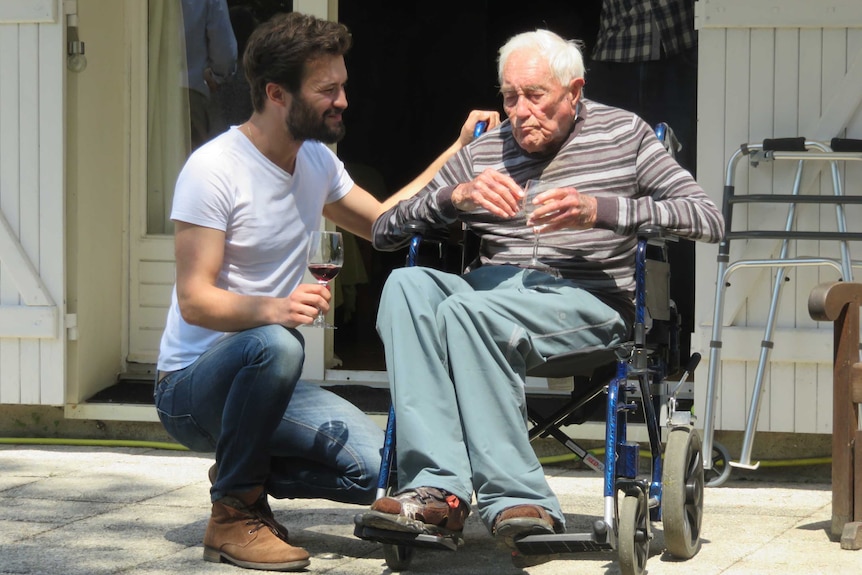 David Goodall sits in a wheelchair in the sunshine, clutching a glass of wine, as grandson Daniel crouches and smiles at him
