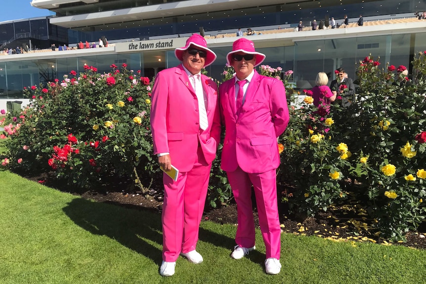 Two men wearing pink suits and hats, stand in front of roses bushes and a race track grandstand.