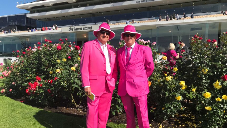 Two men wearing pink suits and hats, stand in front of roses bushes and a race track grandstand.