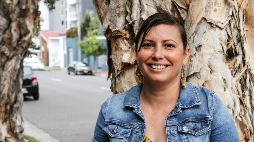 A smiling woman, seemingly in her thirties, standing in front of a tree.