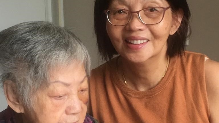 Rebecca Chong stands beside her 86-year-old mum Chan Kai Let,who is seated at a table.
