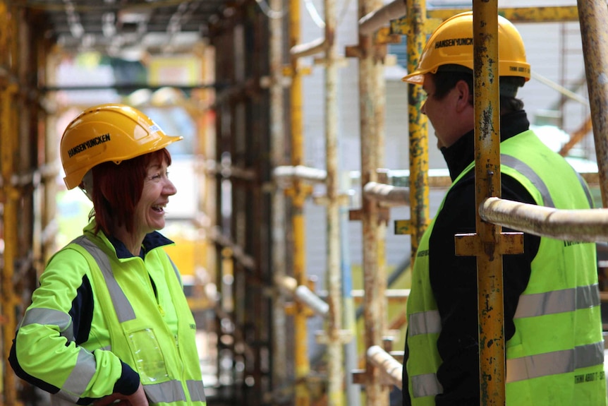 A woman in a hard hat and hi-vis speaks to a man amid scaffolding