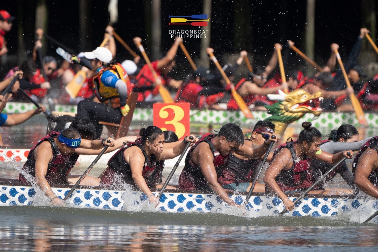 Estudantes universitários em um barco, remando em uma corrida de barcos-dragão em um rio nas Filipinas com outras equipes ao fundo.