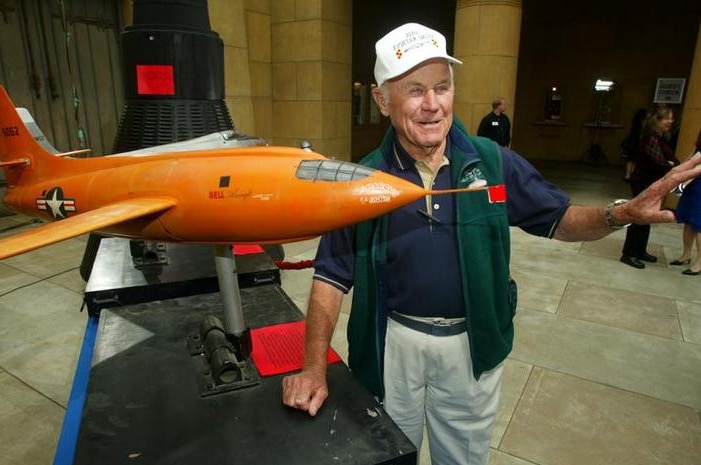 Chuck Yeager stands next to the model of an orange plane and looks to the right of the shot. He holds an arm out.