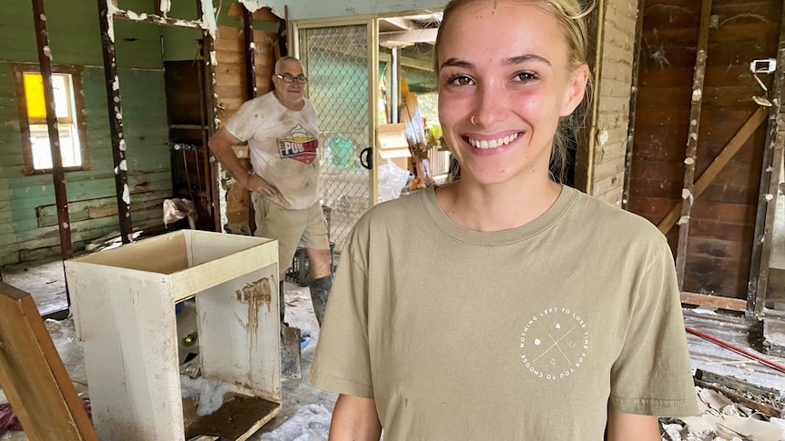 Bianca Pope stands in a damged home, smiling, her dad Gary is in the background.