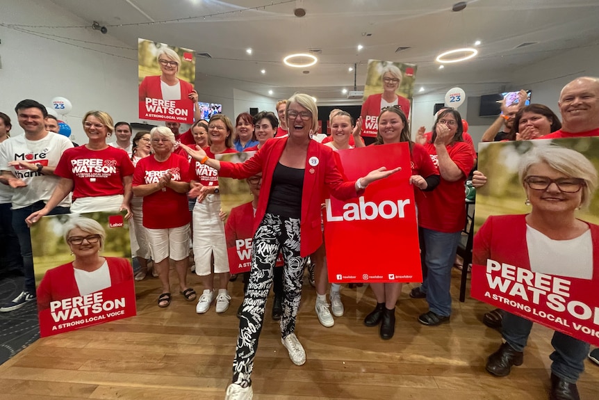 A group of Labor supporters dressed in red stand around a woman with her arms out, celebrating.