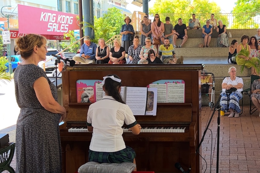 A student plays a piano with her back to the camera while an audience watches in the background .