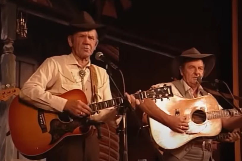 Two older men with guitars standing on stage wearing akrubras, both wear brown pants, cream shirt, one has a vest.