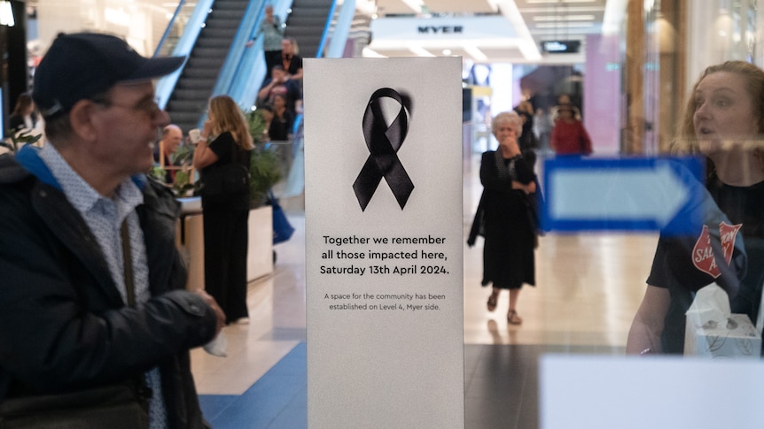 Through the doors of a shopping centre people are seen walking around, there is a sign that says 'together we remember'