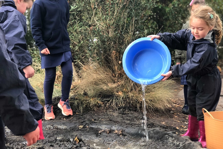 A small child pours water into a mud pit from a blue bucket.