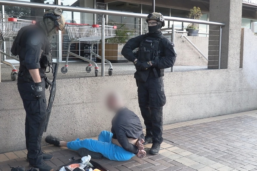 two police stand over a man with a blurred face sitting on the ground
