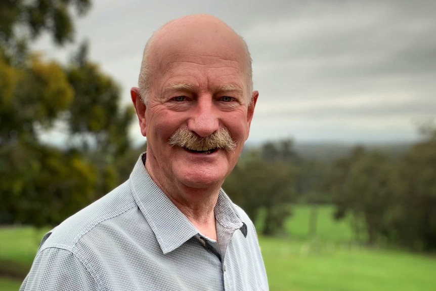 A portrait of a bald man with a mustache smiling as he stands outside on a green hillside.