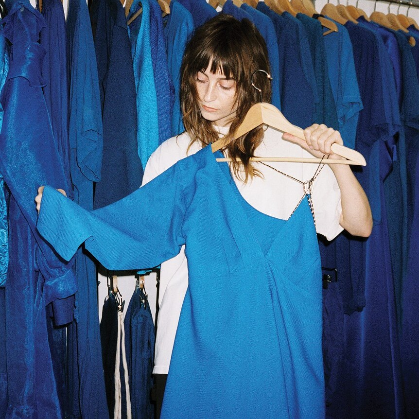 Faye Webster is standing in front a rack of blue dresses, holding one of them in front of her whilst looking down at it. 